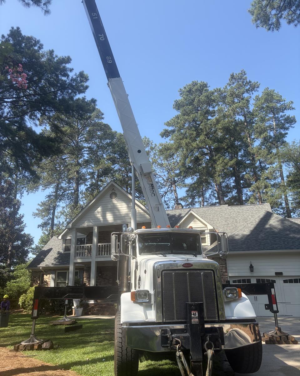 The crane truck set up on outriggers in a driveway, boom extended over the house roof toward tall pines