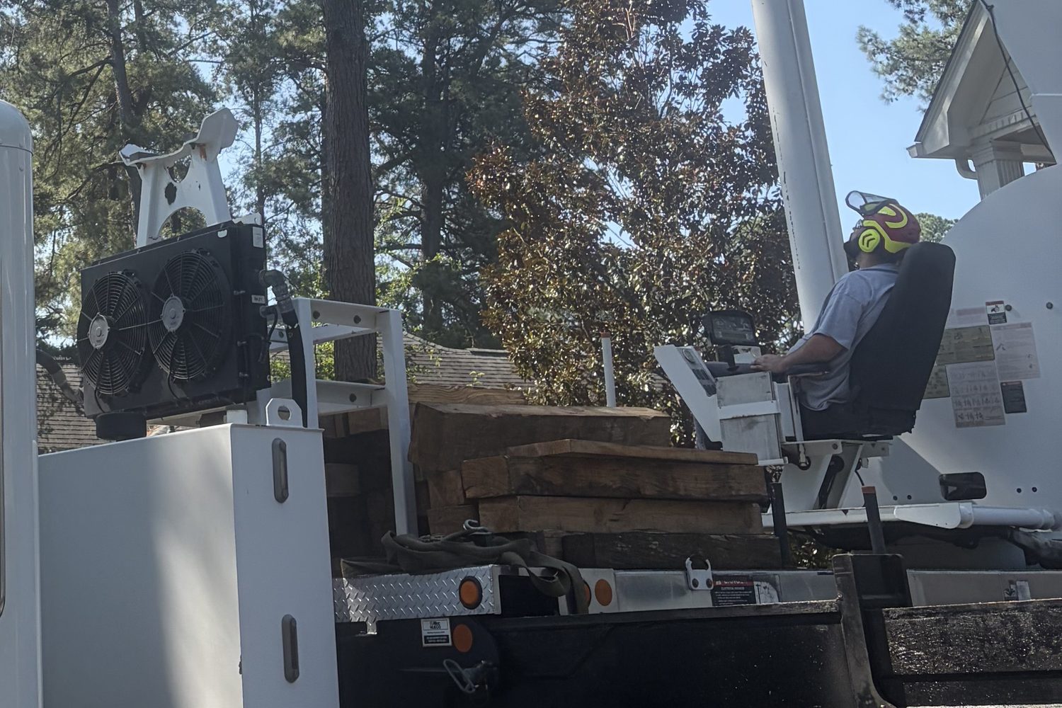 A crane operator in a helmet and ear protection seated at the controls on the back of the truck, with timber mats stacked on the deck beside him and tall pines behind