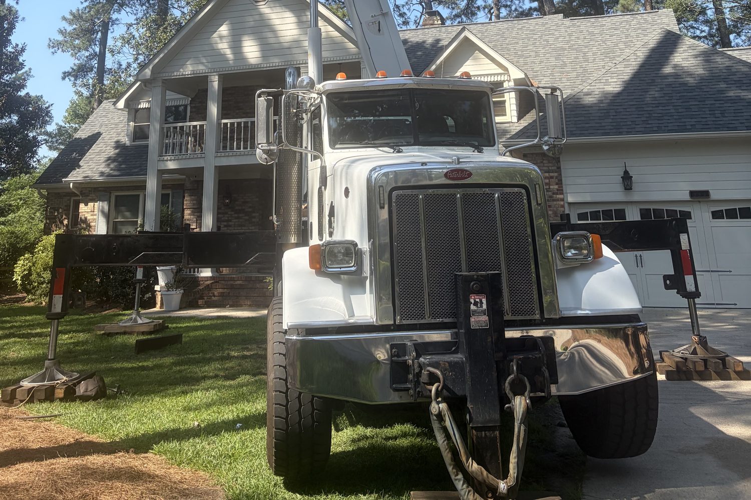 The front of a white crane truck parked along the edge of a driveway, its outrigger legs extended to both sides and resting on stacked timber pads, one on the lawn and one on the concrete