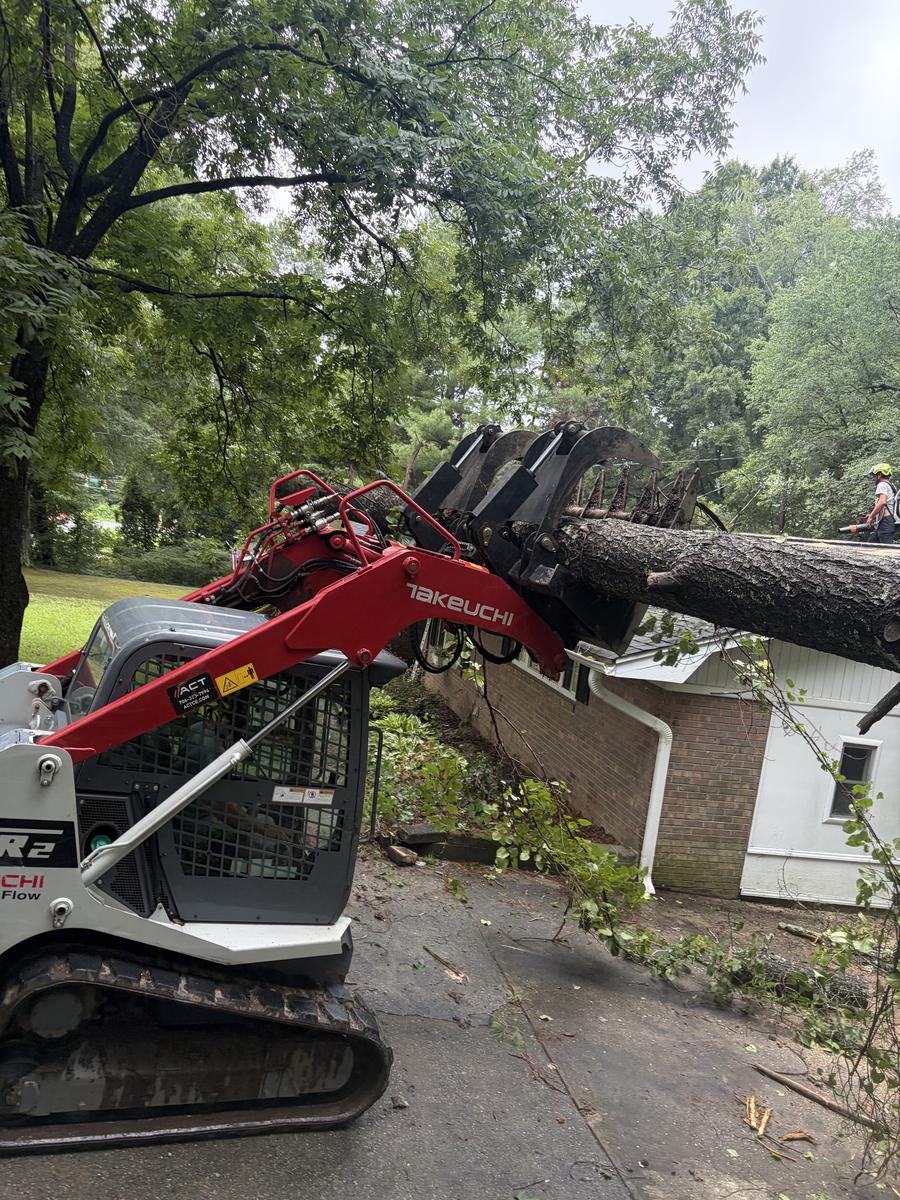 A red tracked grapple machine gripping the fallen trunk at the corner of the house roof